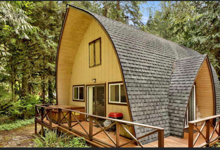 Small yellow cabin with a steep roof and wood deck surrounded by trees.