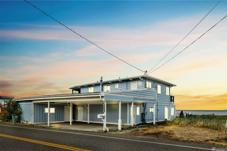 Blue two story home beside a road with a covered porch and water in the distance.