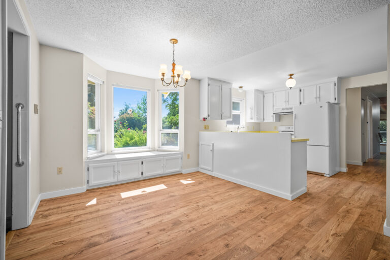 Empty kitchen and dining area with white cabinets, bay windows and wood floors.